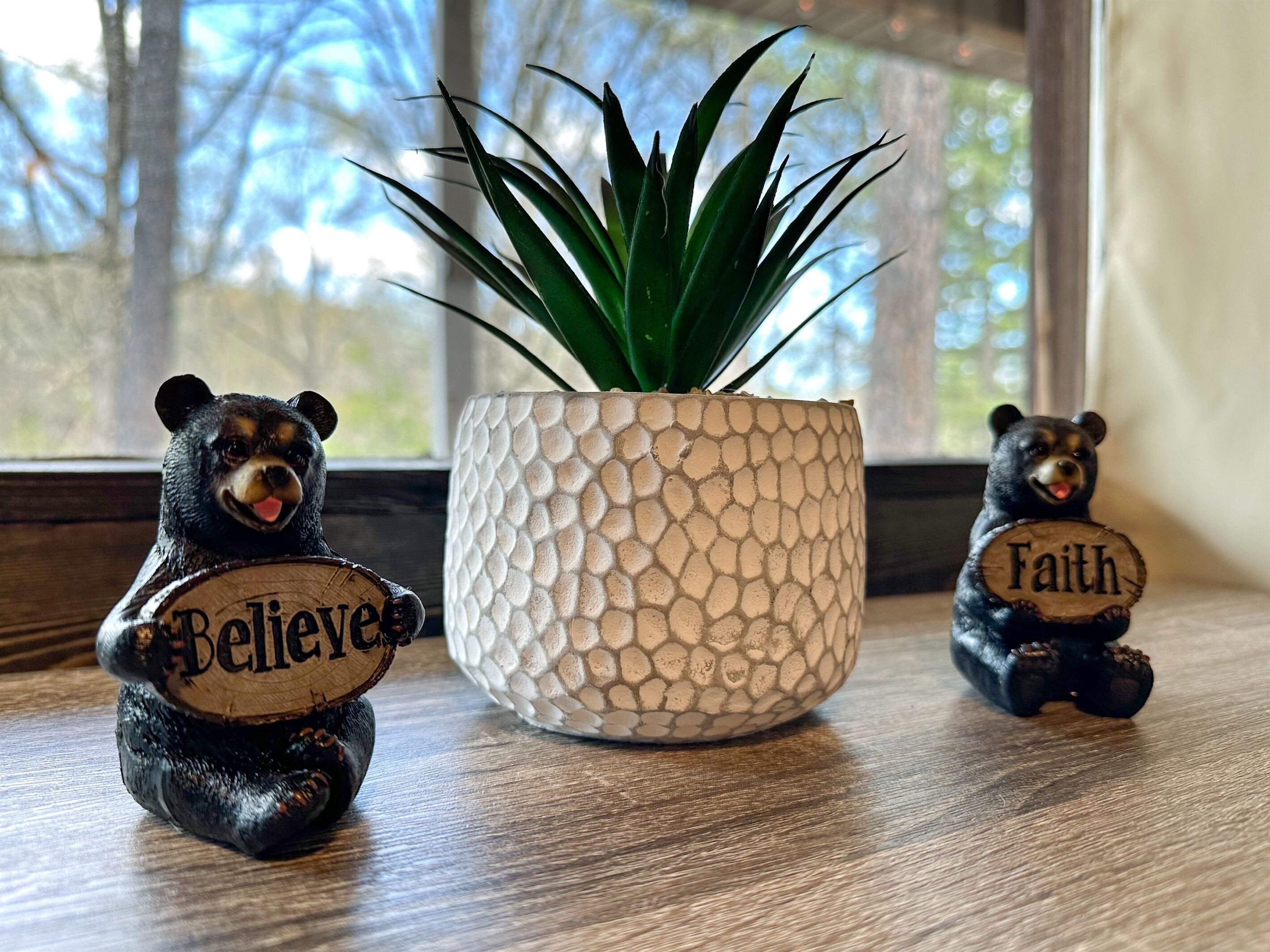Zwei Bärenfiguren halten Schilder mit den Aufschriften 'Believe' und 'Faith'. Neben ihnen steht ein topfgefäß auf einer Holzoberfläche.