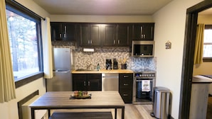A compact kitchen with dark cabinets, a microwave, a stove, and a sink. There's a table with a plate of food and a trash can. - Mama Bear Cabin 2 - Community pool and hottub. (Branson)