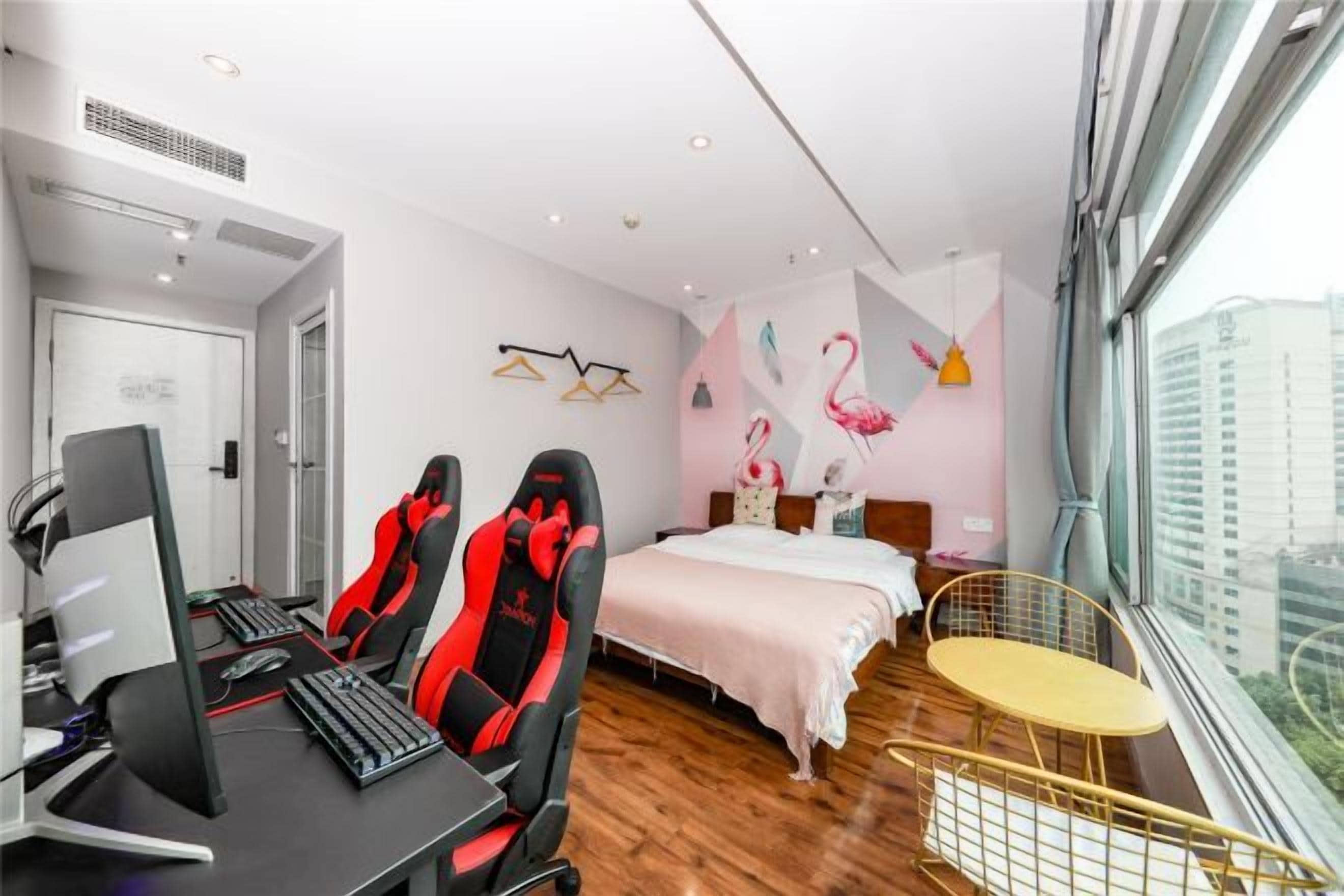 A hotel room with a bed, a desk with a computer, two gaming chairs, a small round table, and a window with a view of buildings.