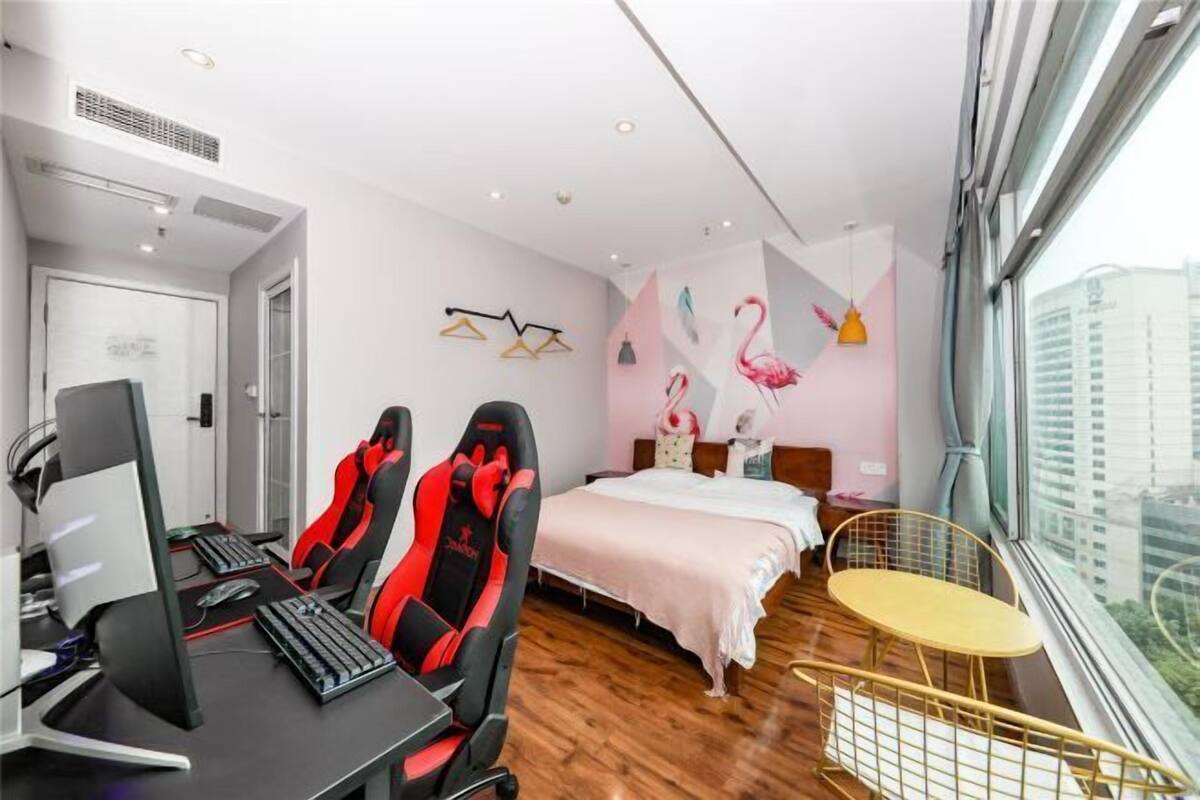 A modern hotel room with a desk, two gaming chairs, a bed, a small round table, and a large window offering a city view.