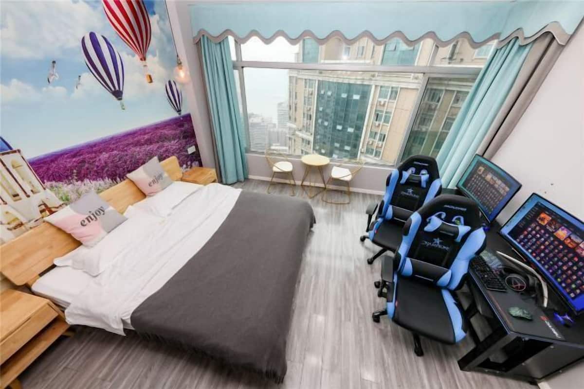 A hotel room with a bed, a desk with a computer, a gaming chair, and a view of the city through large windows.
