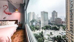 A bedroom with a city view, a bed with a pink bedspread, a wooden floor, and a wall mural of a flamingo.