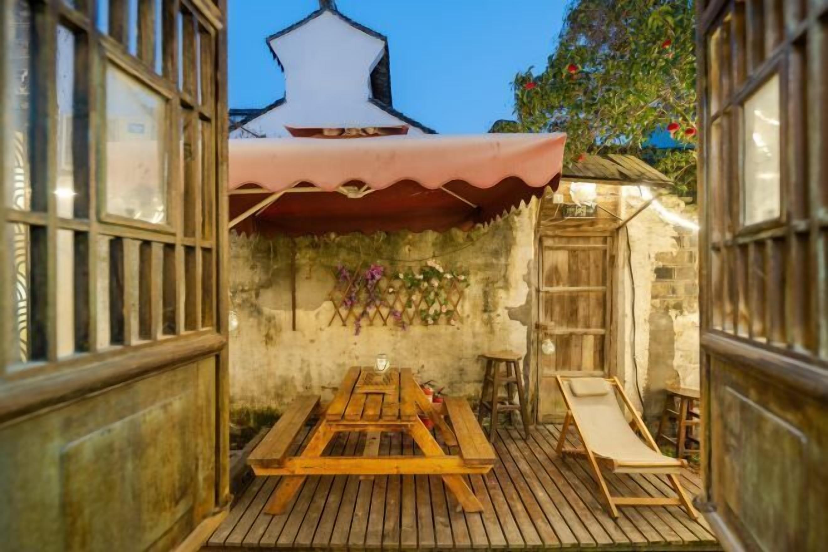 A wooden deck with a picnic table and a lounge chair, surrounded by stone walls and a white building with a pointed roof.