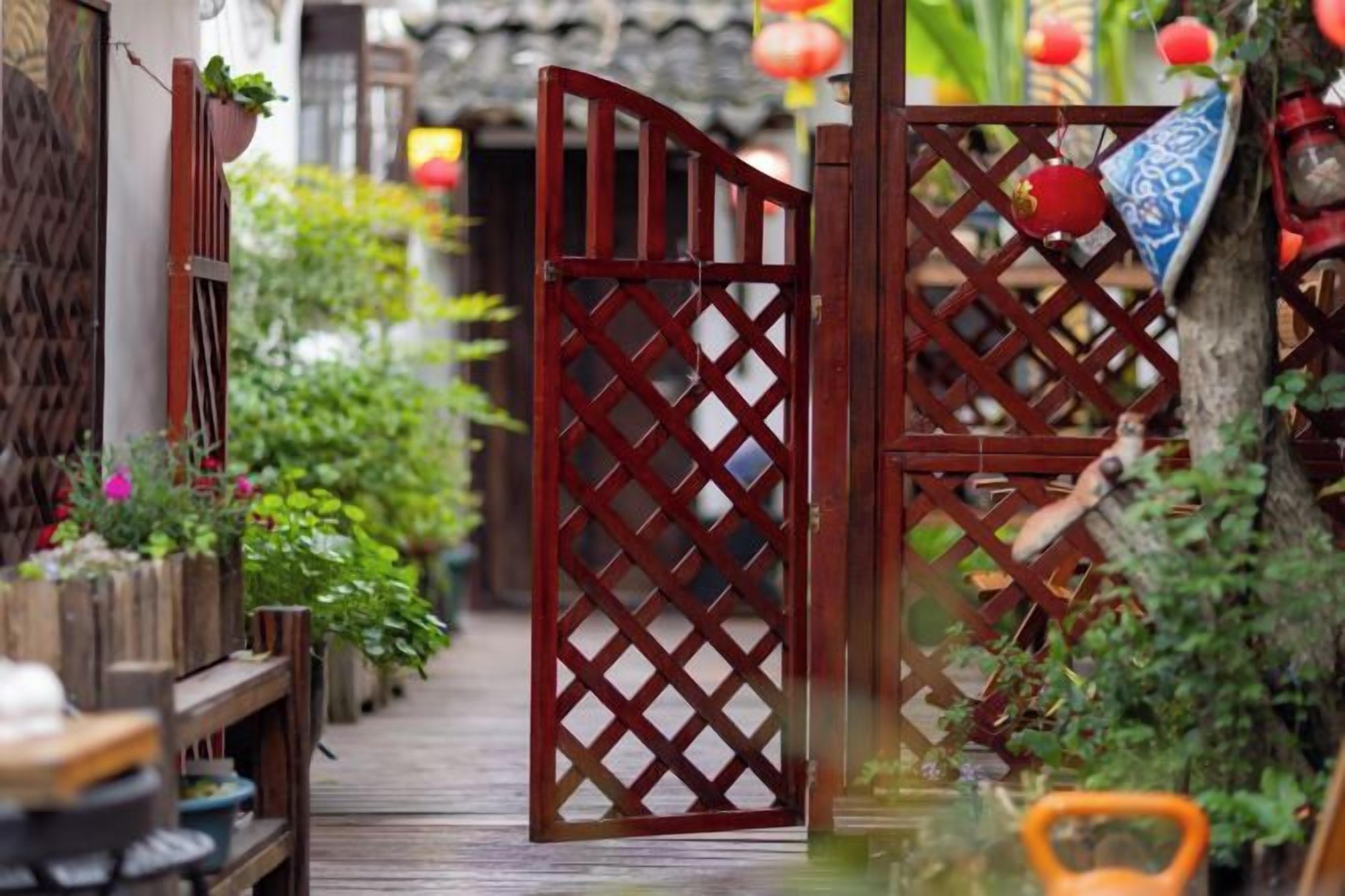 Una reja de madera tradicional con diseño de celosía, que da acceso a un sendero bordeado por plantas en macetas y un banco de madera.