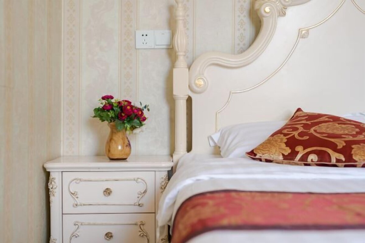 Een net opgemaakt bed met een wit hoofdeinde en een decoratief kussen. Een klein nachtkastje met een bloevas is zichtbaar.
