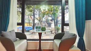 Ein Zimmer mit zwei Sofas, die einem kleinen Tisch mit einem grünen Gegenstand gegenüberliegen, großen Fenstern mit Vorhängen und Blick ins Freie.
