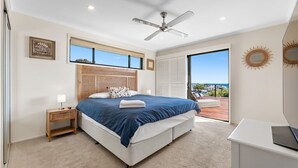 Une chambre avec un grand lit, un ventilateur de plafond, une table de chevet, une lampe, une vue sur l’océan et un miroir en forme de soleil.