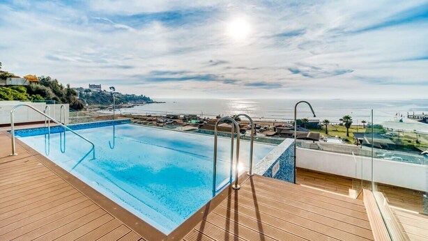 A pool with a wooden deck and glass railings, overlooking a beach and ocean. - Miramar Deluxe con Vista al Mar (Concón)