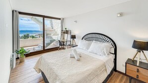 A bedroom with a bed, a nightstand, a lamp, a potted plant, and a view of the sea through a large window. - Miramar Deluxe con Vista al Mar (Concón)