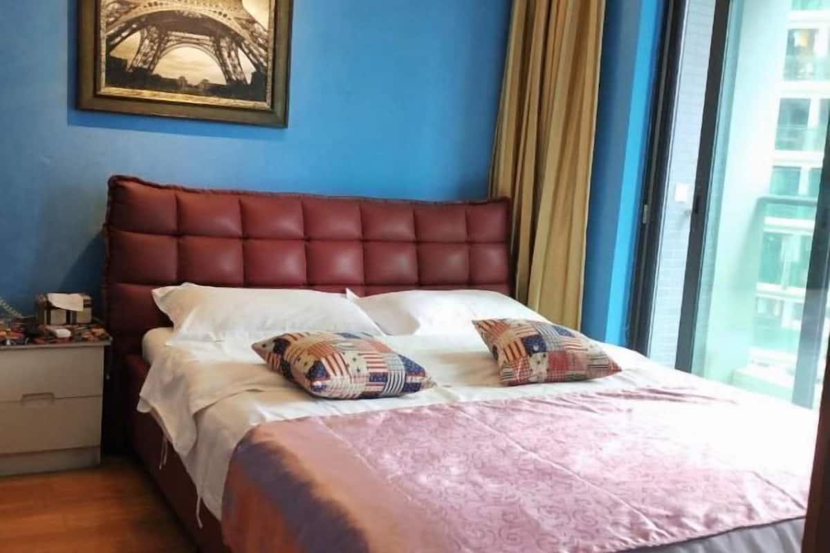 Un dormitorio con cama, almohadas, una mesita de noche, un cuadro enmarcado en la pared y una ventana con cortinas.