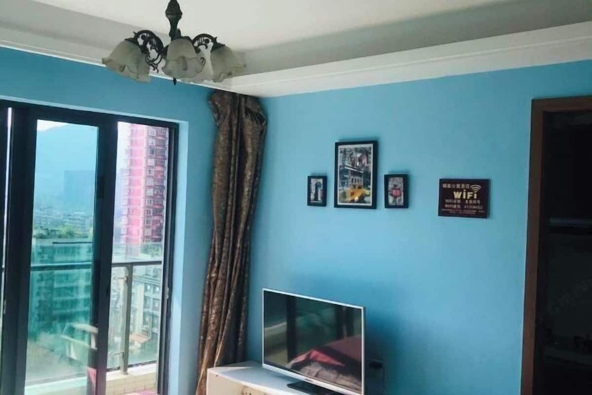Una habitación con un ventanal grande, un televisor de pantalla plana sobre un soporte, una lámpara de techo y un cartel en la pared que indica acceso a WiFi.