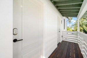 Une porte blanche dotée d’un clavier pour ouvrir et d’une poignée noire, qui donne sur une terrasse en bois avec des balustrades blanches et une vue sur des palmiers.