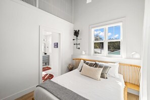 Une chambre avec un grand lit, une tête de lit en bois, une fenêtre donnant sur un beau paysage et un mur blanc agrémenté d’un élément décoratif noir.