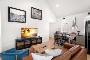 Ein modernes Wohnzimmer mit Couchtisch aus Holz, blauem Sessel und braunem Ledersofa. Es gibt einen Essbereich mit Holztisch und Stühlen sowie eine Küche mit schwarzem Kühlschrank und Edelstahl-Dunstabzugshaube.