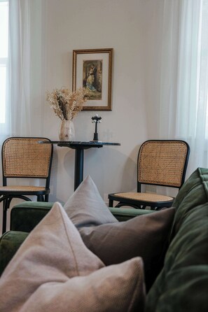 Una habitación con dos sillones de mimbre, una mesita con un jarrón y un marco de fotos, y un sofá verde con un cojín.
