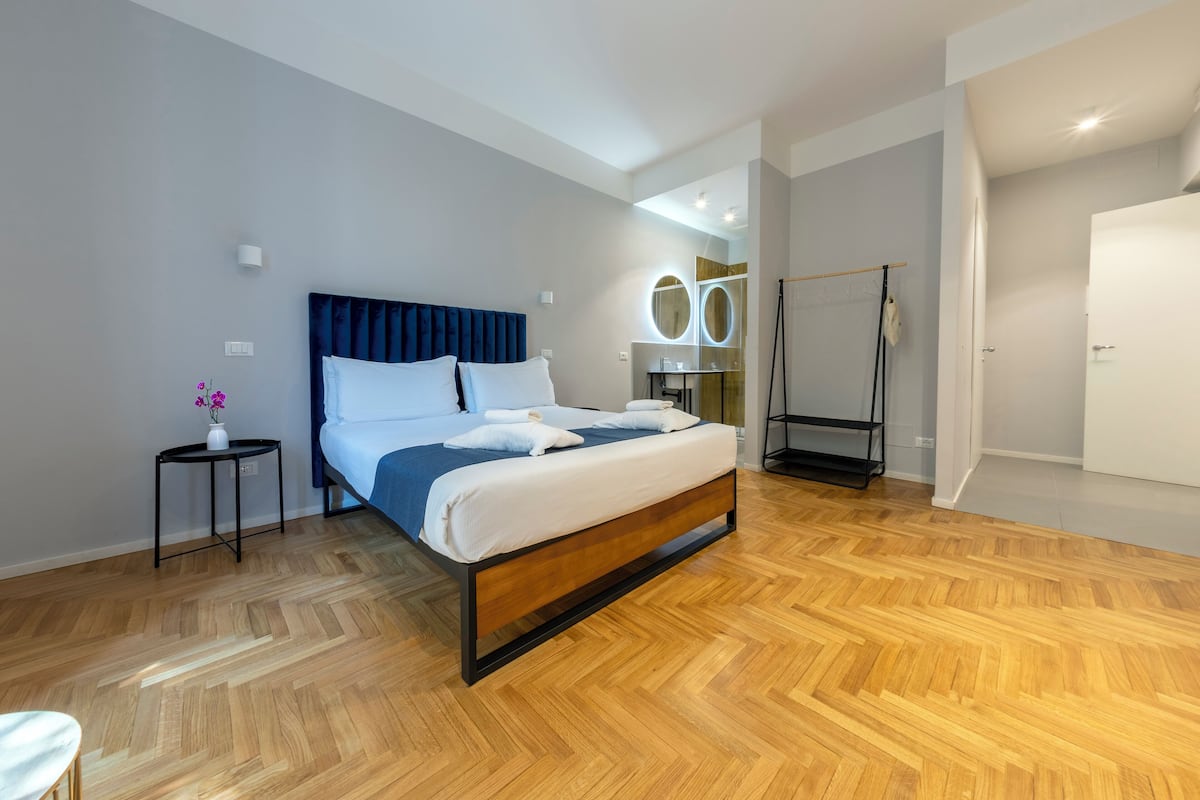 A modern hotel room with a large bed, wooden flooring, a small side table with a vase, and a mirror with circular lights.