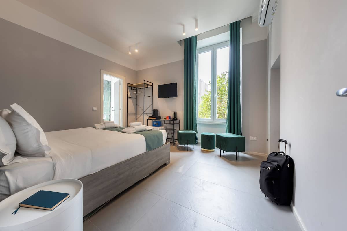 A modern hotel room with a large bed, a desk, two green ottomans, a black suitcase, and a flat-screen TV on the wall.