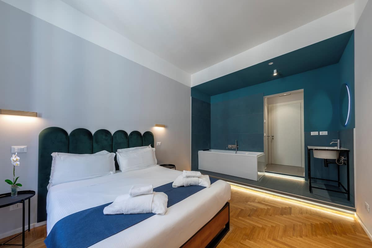 A modern hotel room with a large bed, a bedside table with a lamp, a bathroom with a mirror, and a sink.