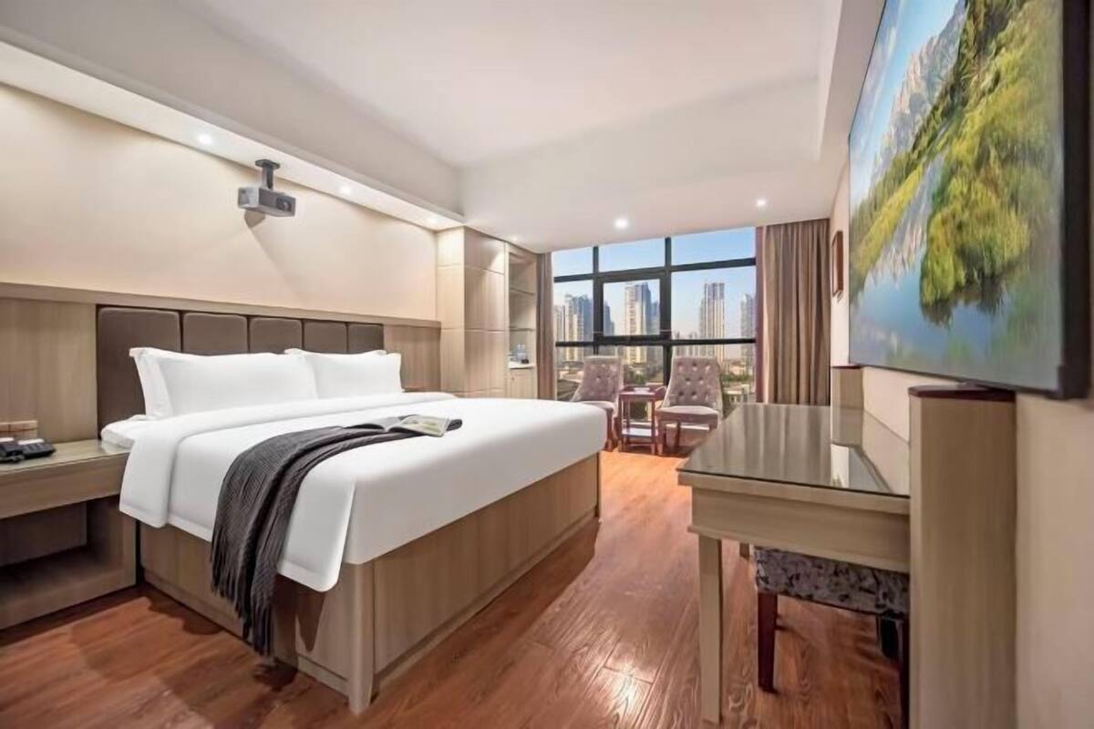 A hotel room with a large bed, a desk, a chair, and a large window offering a city view.