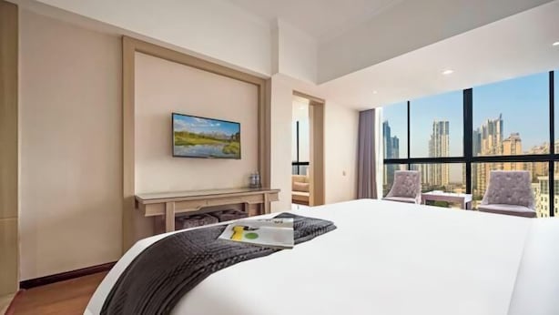 A hotel room with a large bed, a painting on the wall, a desk, and a view of the city through large windows. - Dingyi Junyue Hotel (Changde)