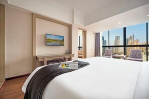 A hotel room with a large bed, a painting on the wall, a desk, and a view of the city through large windows. - Dingyi Junyue Hotel (Changde)