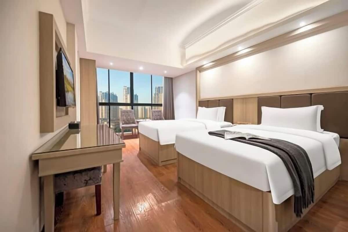 A hotel room with two beds, a desk, and a large window offering a city view.