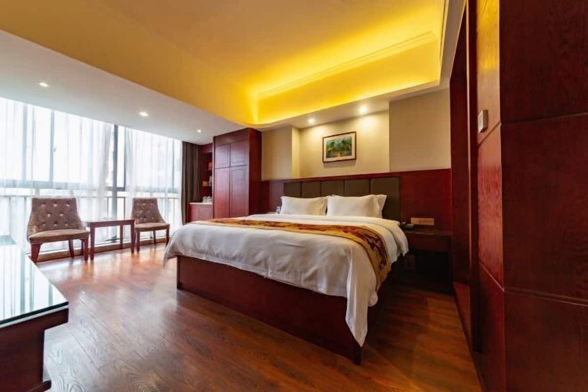 A hotel room with a large bed, wooden flooring, a desk, two chairs, and a large window.