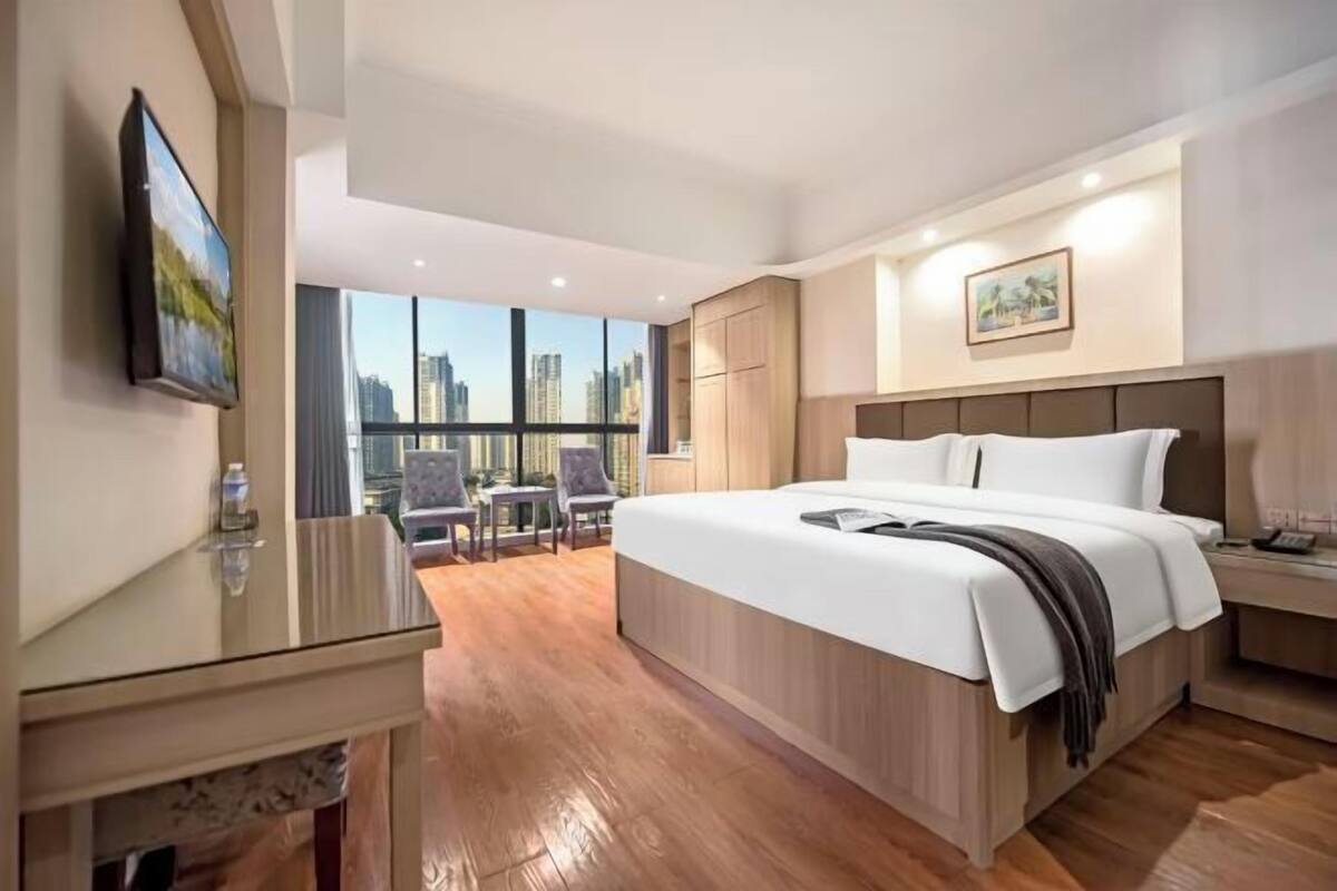 A hotel room with a large bed, a desk with a chair, a small table with chairs, and a large window offering a city view.