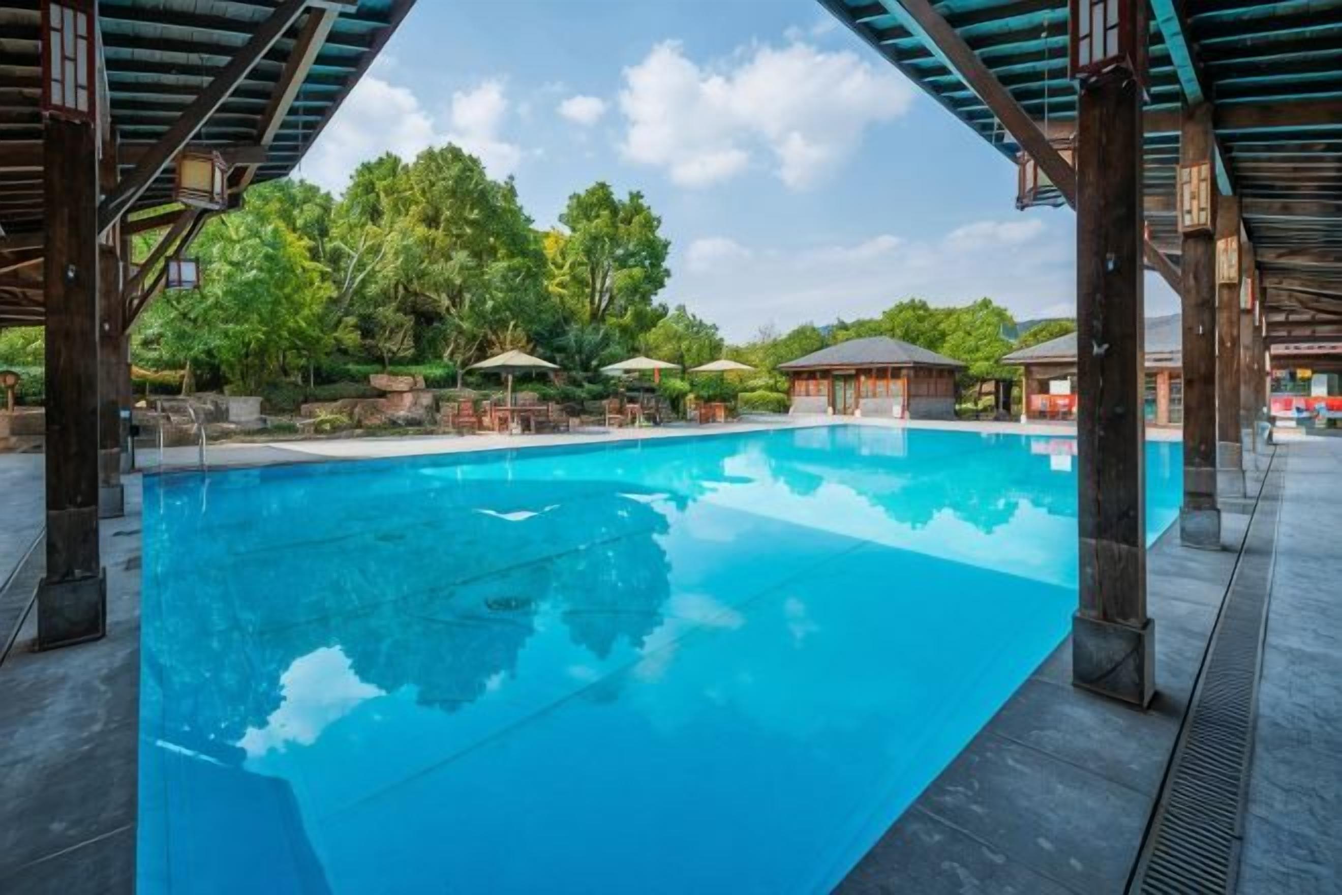 A clear blue swimming pool with a covered area, surrounded by trees and outdoor seating.