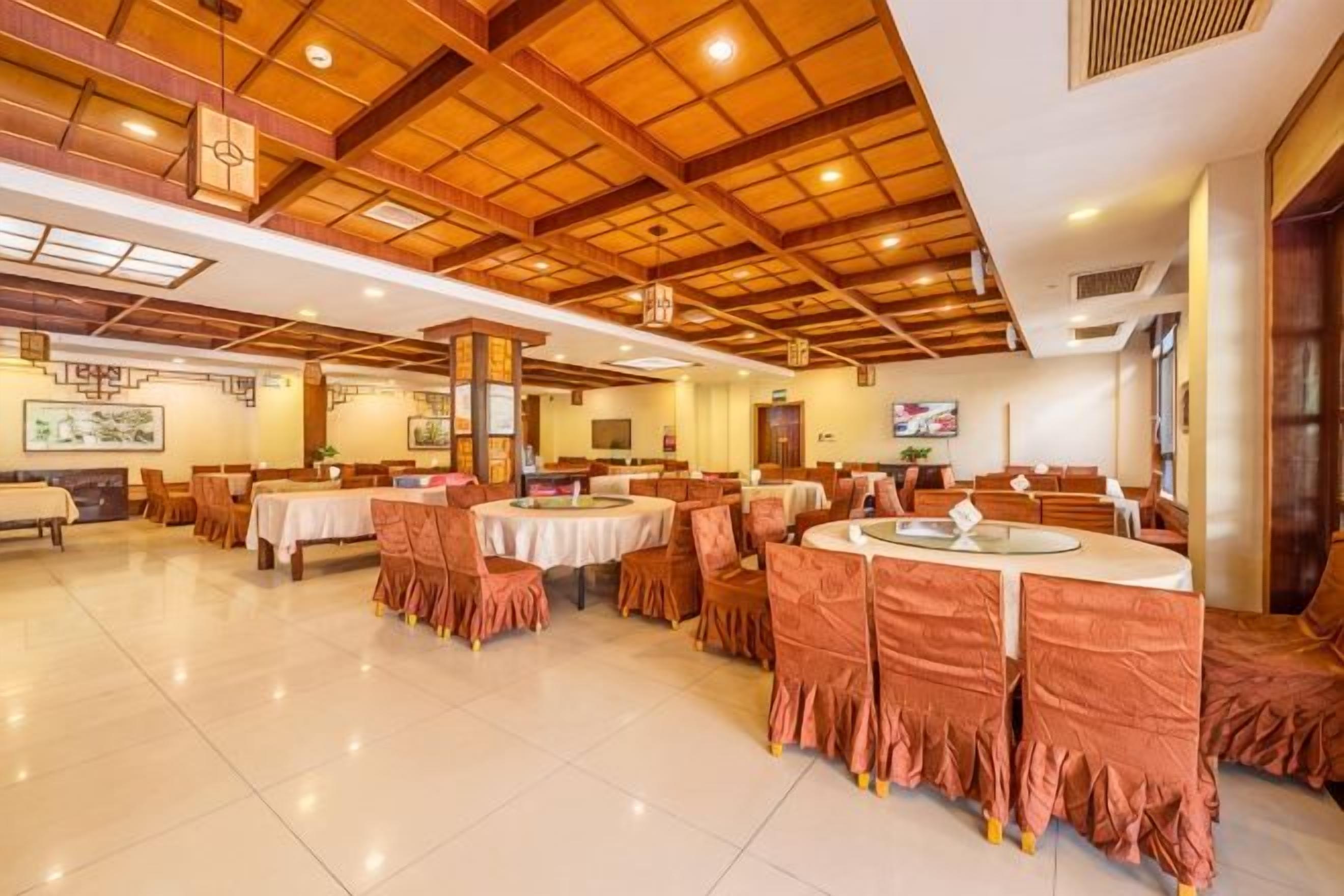 A spacious dining area with multiple round tables, each surrounded by chairs. The room features a high wooden ceiling with recessed lighting and a tiled floor.