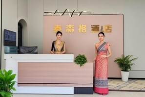 A reception desk with a sign above it, two individuals standing beside it, and a potted plant to the right. - Taisenge Hotel (Jinghong)