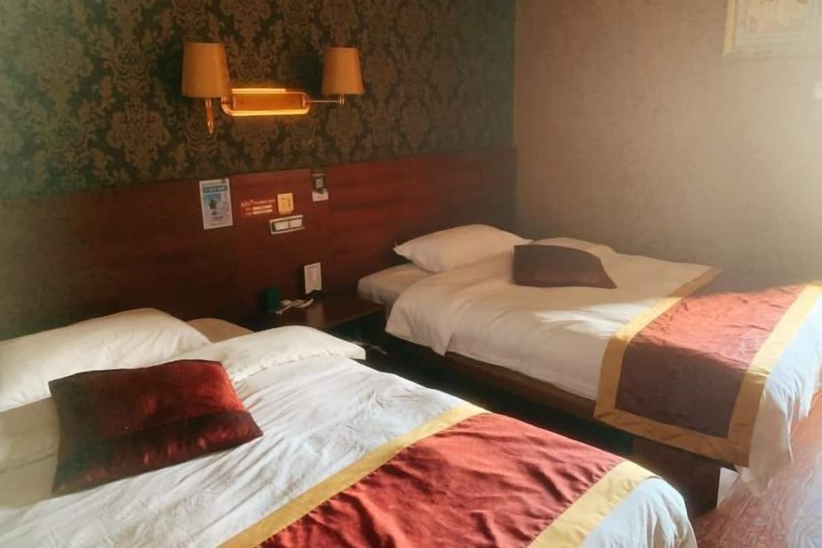 Une chambre d’hôtel avec trois lits, chacun doté de draps blancs et d’un élément décoratif rouge. Des lampes murales sont fixées sur le côté gauche de la tête de lit.