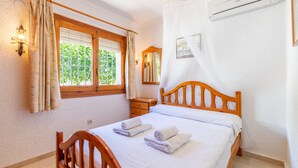 A bedroom with a bed, a nightstand, a window with curtains, a mirror, and an air conditioning unit. - Villa 'Feliz ' with Private Pool, Wi-Fi and Air Conditioning (Calpe)