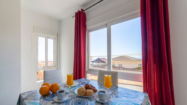Una mesa de desayuno con naranjas, cruasanes y jugo, una silla blanca y una ventana con cortinas rojas.