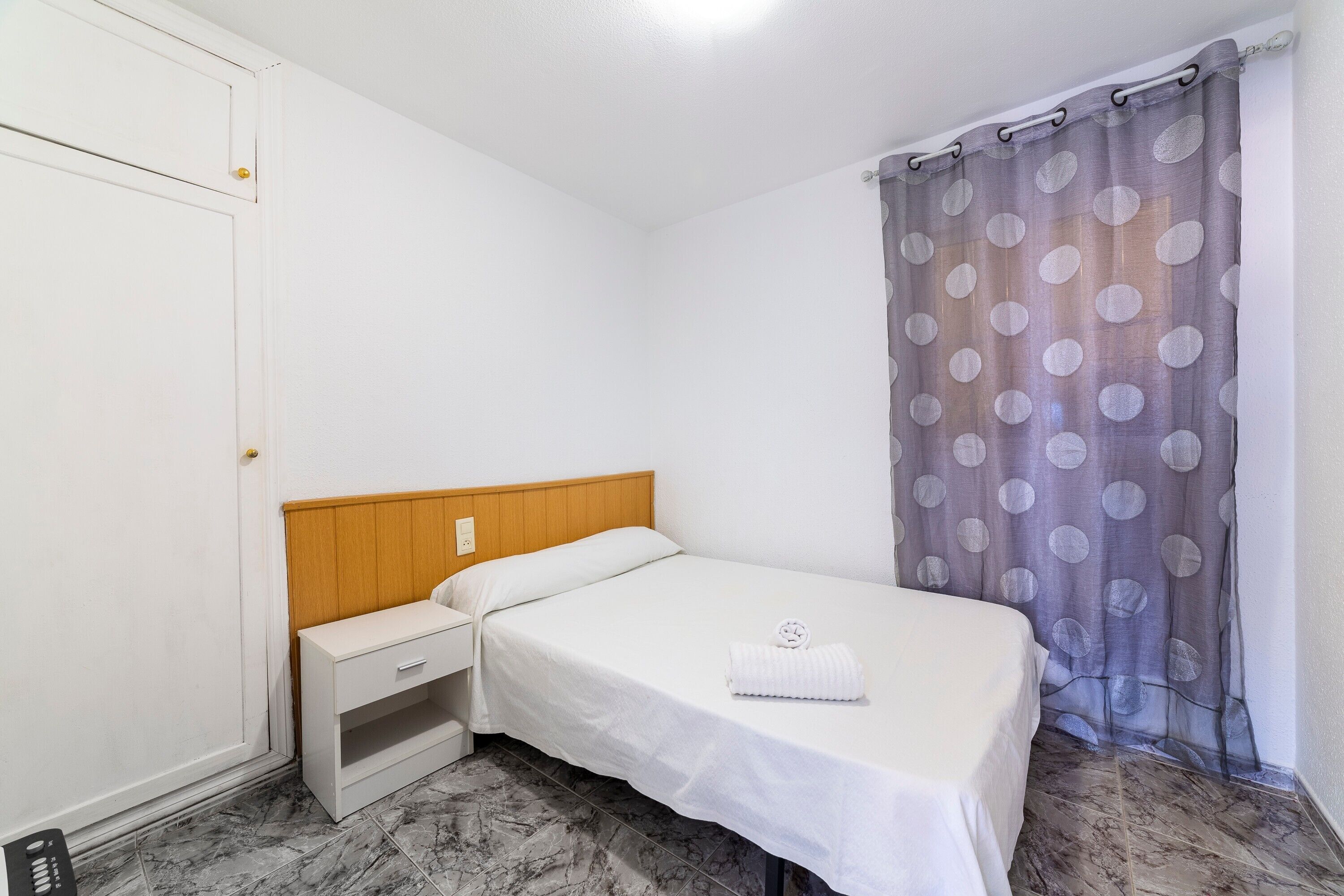 Una cama individual con cabecera de madera, una mesita de noche y una cortina gris con estampado de lunares.