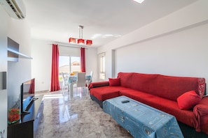 A modern living room with a red sofa, a blue ottoman, a flat-screen TV, and a dining area with a table and chairs.