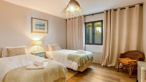 A bedroom with two beds, a bedside table with a lamp, a wicker chair, and a window with curtains. - Apartment 'Referta Apartamento T2' with Sea View, Shared Garden and Wi-Fi (Prazeres)