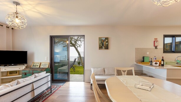 A modern living room with a flat-screen TV, sofa, dining table, and a view through a glass door to an outdoor area. - Apartment 'Referta Apartamento T2' with Sea View, Shared Garden and Wi-Fi (Prazeres)