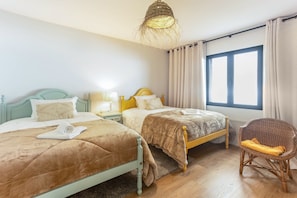 A bedroom with two beds, a wicker chair, a window with curtains, and a hanging light fixture.