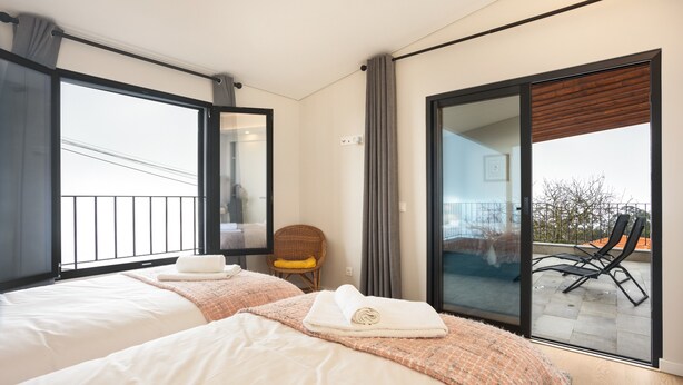 A hotel room with two beds, a wicker chair, a balcony with chairs, and a sliding glass door. - Holiday Home 'Referta House' with Sea View, Private Garden and Wi-Fi (Prazeres)