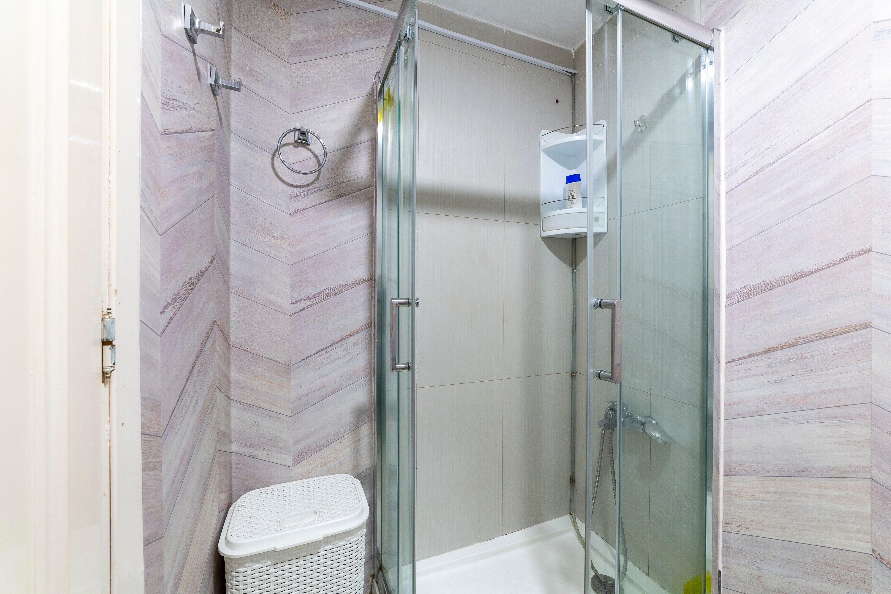 Box doccia in vetro con pavimento piastrellato di bianco, un cesto bianco e porta doccia in vetro.