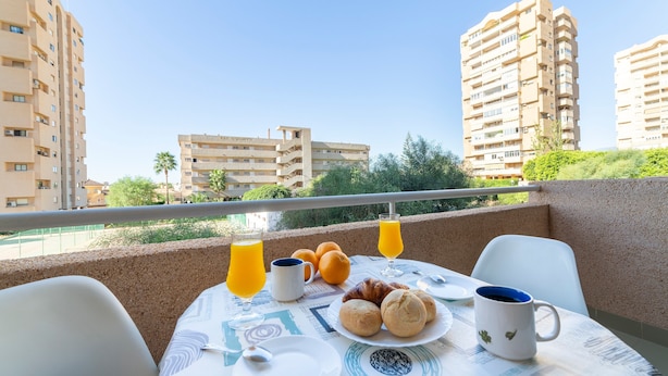 Balcón con productos para el desayuno, como cruasanes, jugo de naranja y una taza de café, con vistas a edificios altos y zonas verdes.