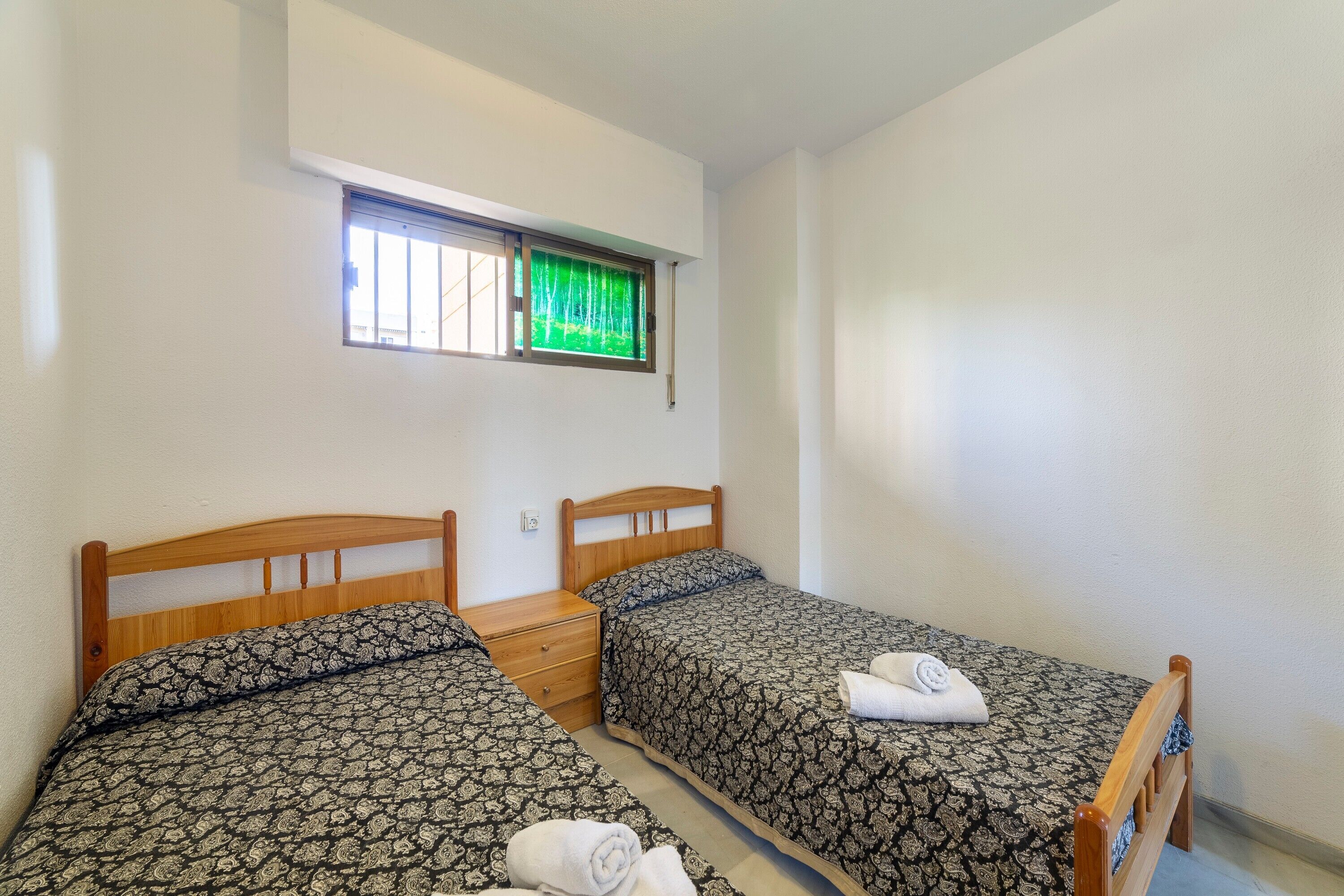 Ein Zimmer mit zwei Einzelbetten, einem kleinen Nachttisch, einem Fenster mit Jalousien und einem an der Wand montierten Lichtschalter.
