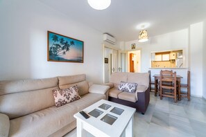 Un salon avec un canapé, une table basse sur laquelle se trouve une télécommande, et un tableau représentant des palmiers.