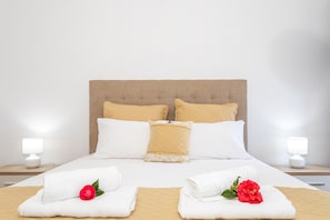 A neatly made bed with white and beige bedding, two white lamps on nightstands, and a red rose on each side.