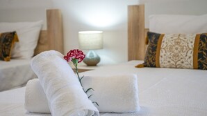 A neatly made bed with a rolled white towel and a single red flower, a bedside table with a vase, and a patterned pillow.