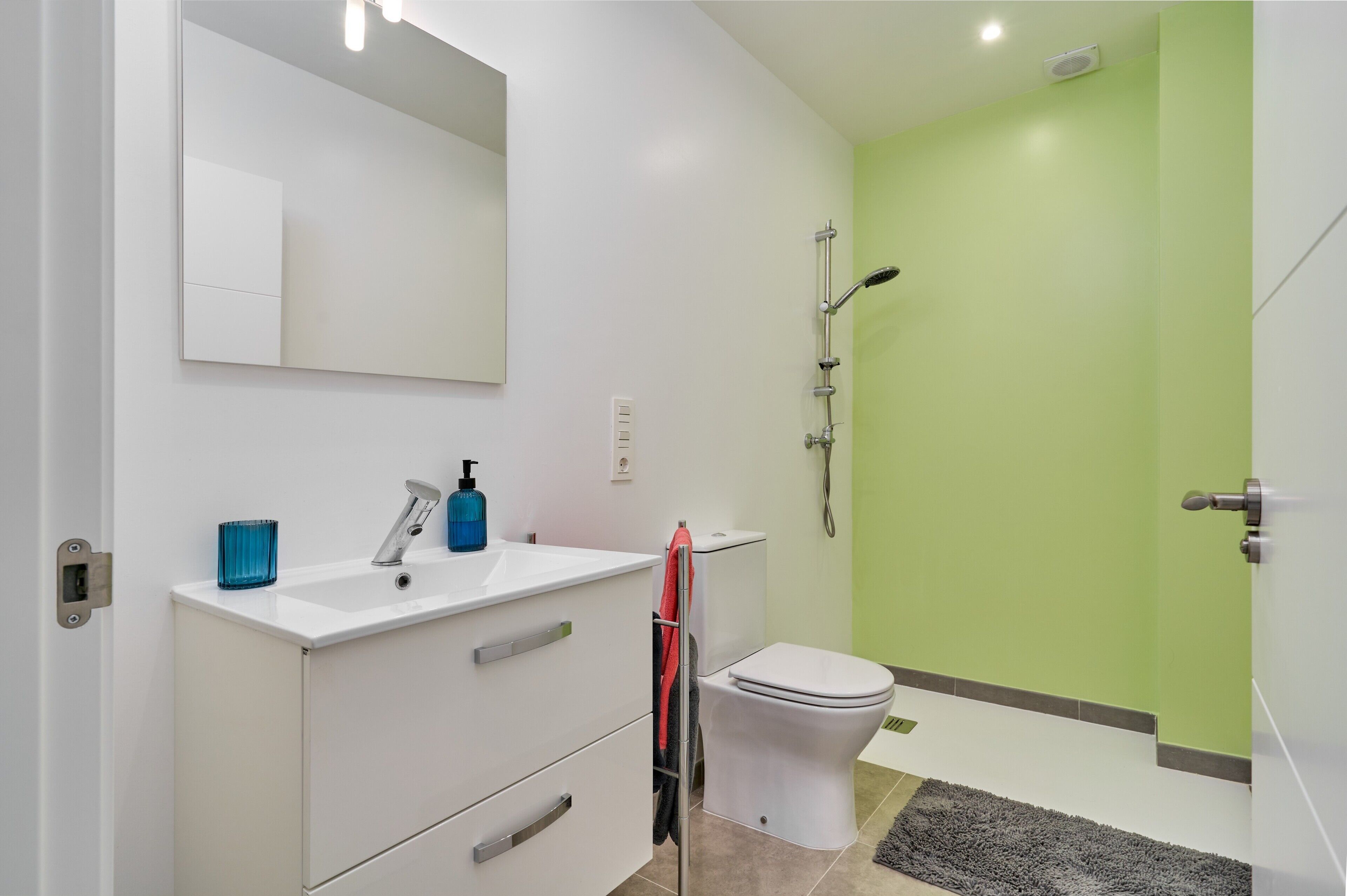 Ein modernes Badezimmer mit Waschbecken, Spiegel und Toilette in Weiß, einer Dusche mit grüner Wand und einem kleinen Teppich auf dem Boden.