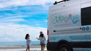 Een strandscène met een witte bus, genaamd 'Campomare on Wheels', geparkeerd in het zand.