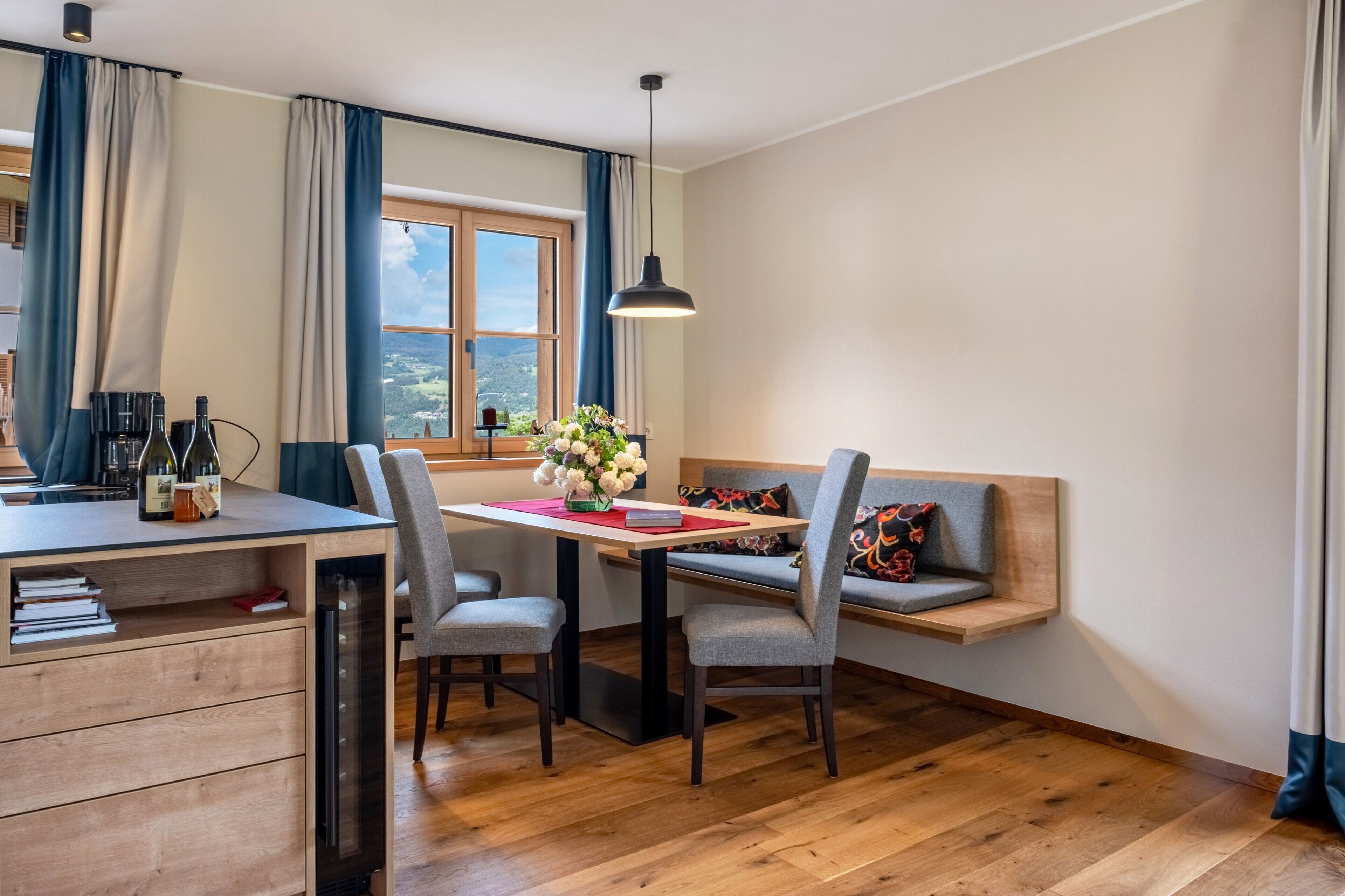 A modern room with a wooden desk, chairs, a bench, a window with curtains, and a hanging light fixture.
