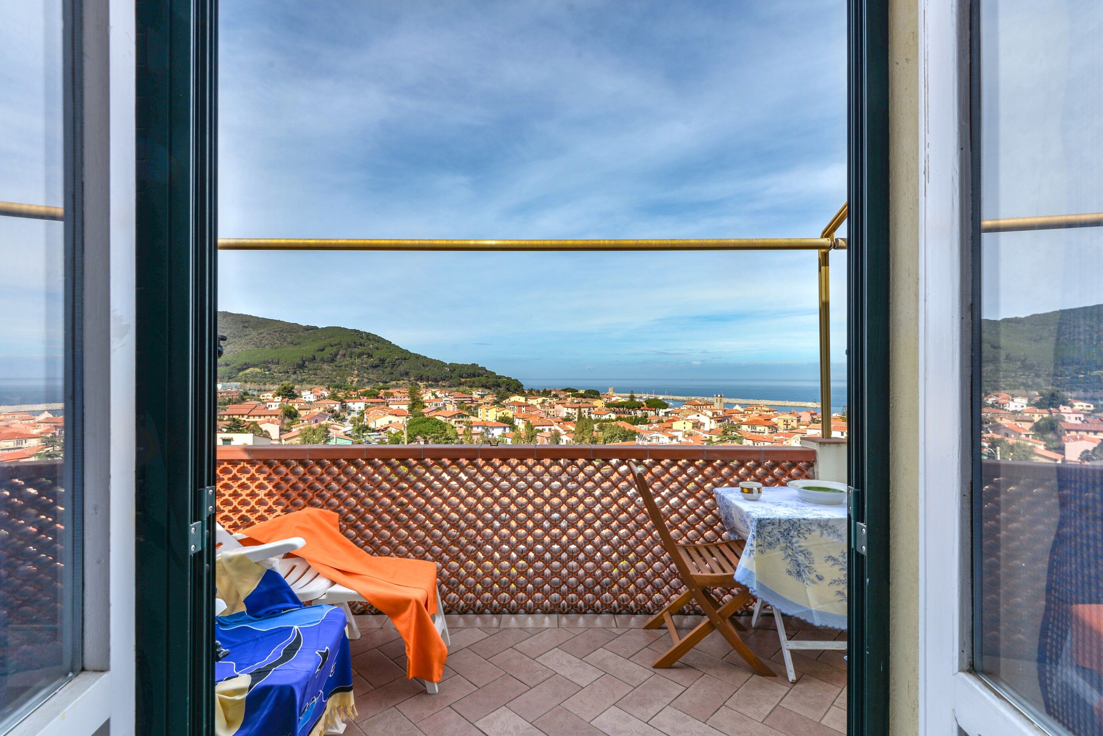Un balcón con vistas a un pueblo costero, una mesa con mantel y una silla.